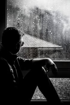 一个戴着太阳镜的男人，剪影映在雨中的窗前，若有所思地坐着