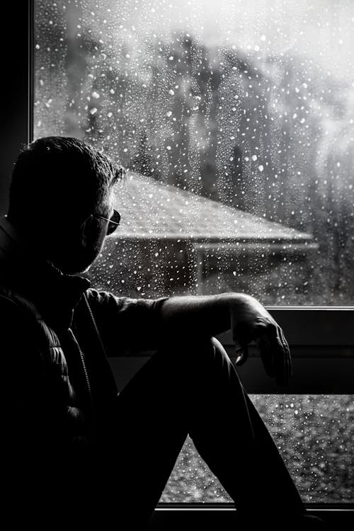 Grayscale Photo of Man Sitting Beside Window 