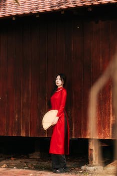 Elegant woman in red Ao Dai holding bamboo fan against a wooden backdrop.