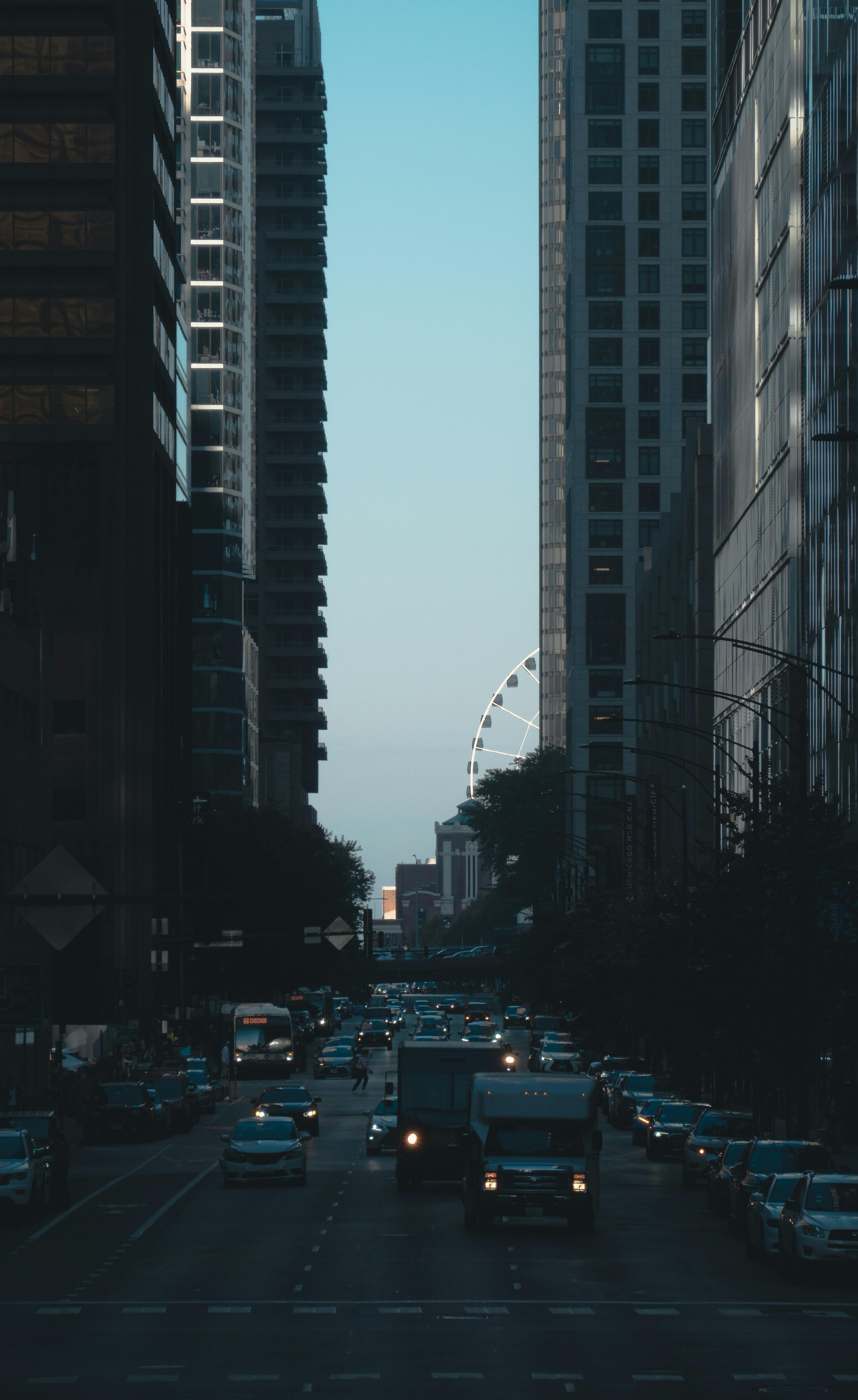 City street with tall buildings, traffic, and a distant Ferris wheel at twilight.