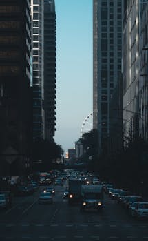 City street with tall buildings, traffic, and a distant Ferris wheel at twilight.