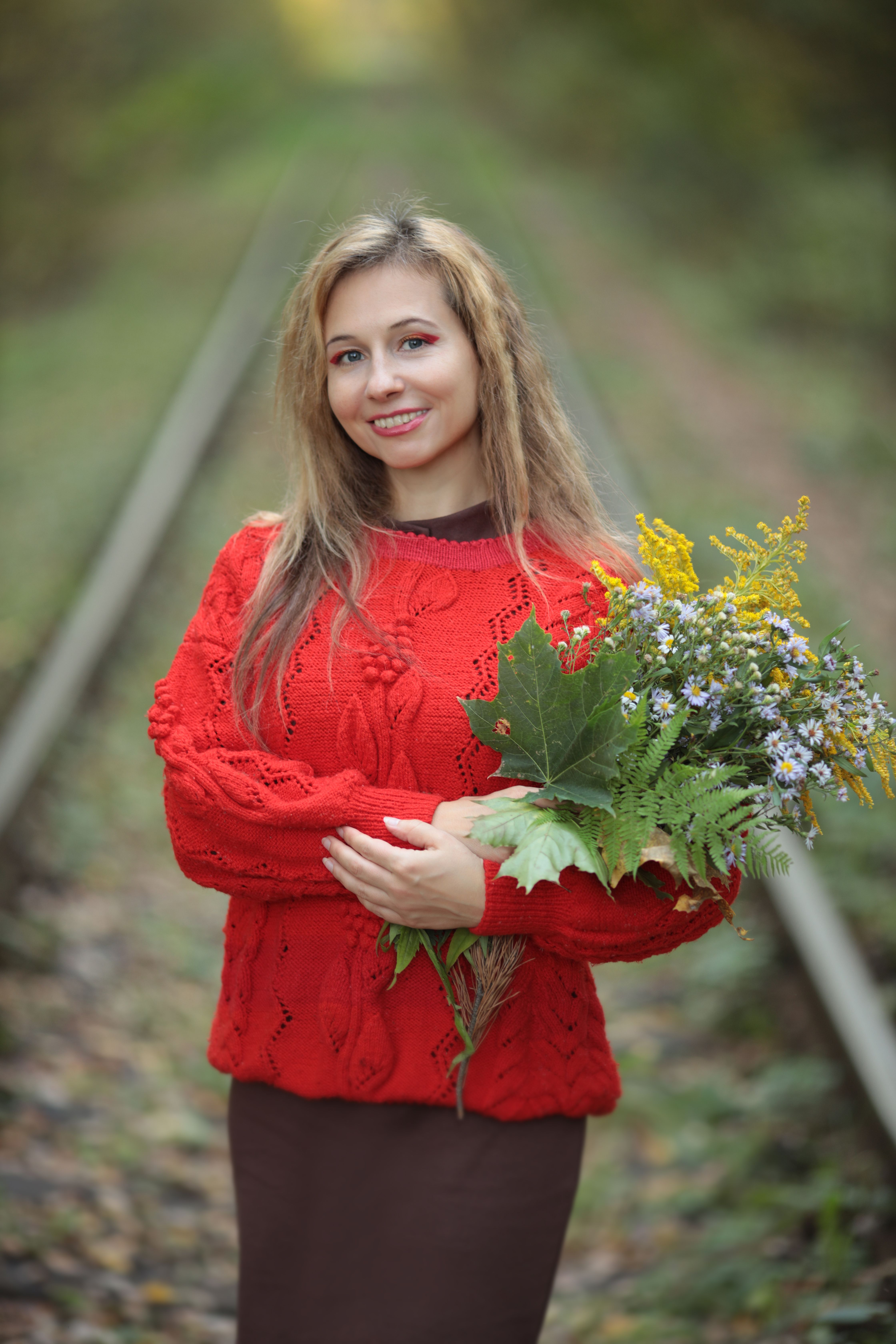 A woman in a red sweater smiles while holding a wildflower bouquet on railway tracks.