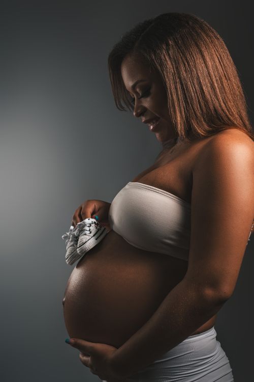 Free Pregnant woman in a studio, hand on belly holding baby shoes, side profile portrait. Stock Photo