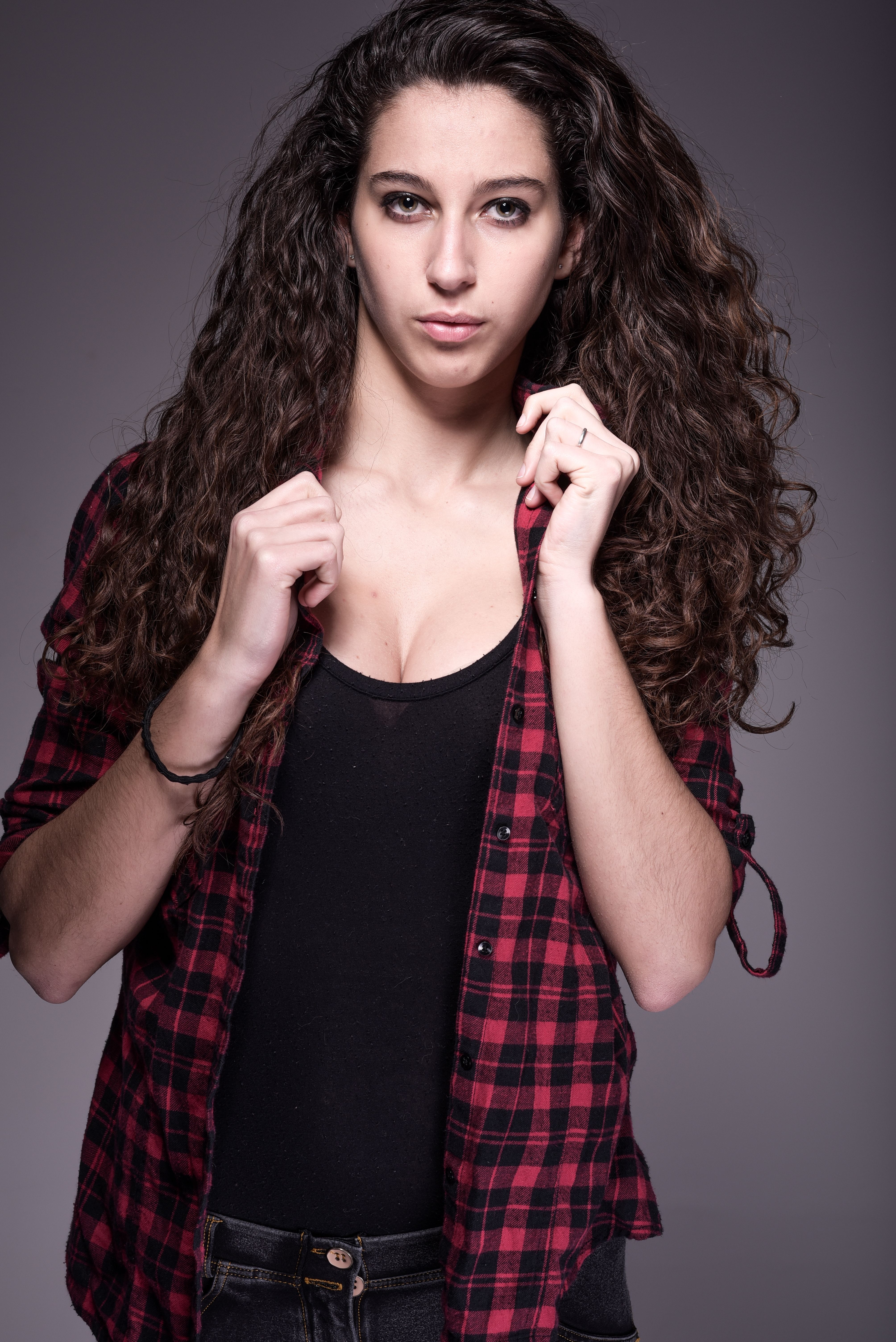 Confident woman with long curly hair in a red plaid shirt, studio portrait.