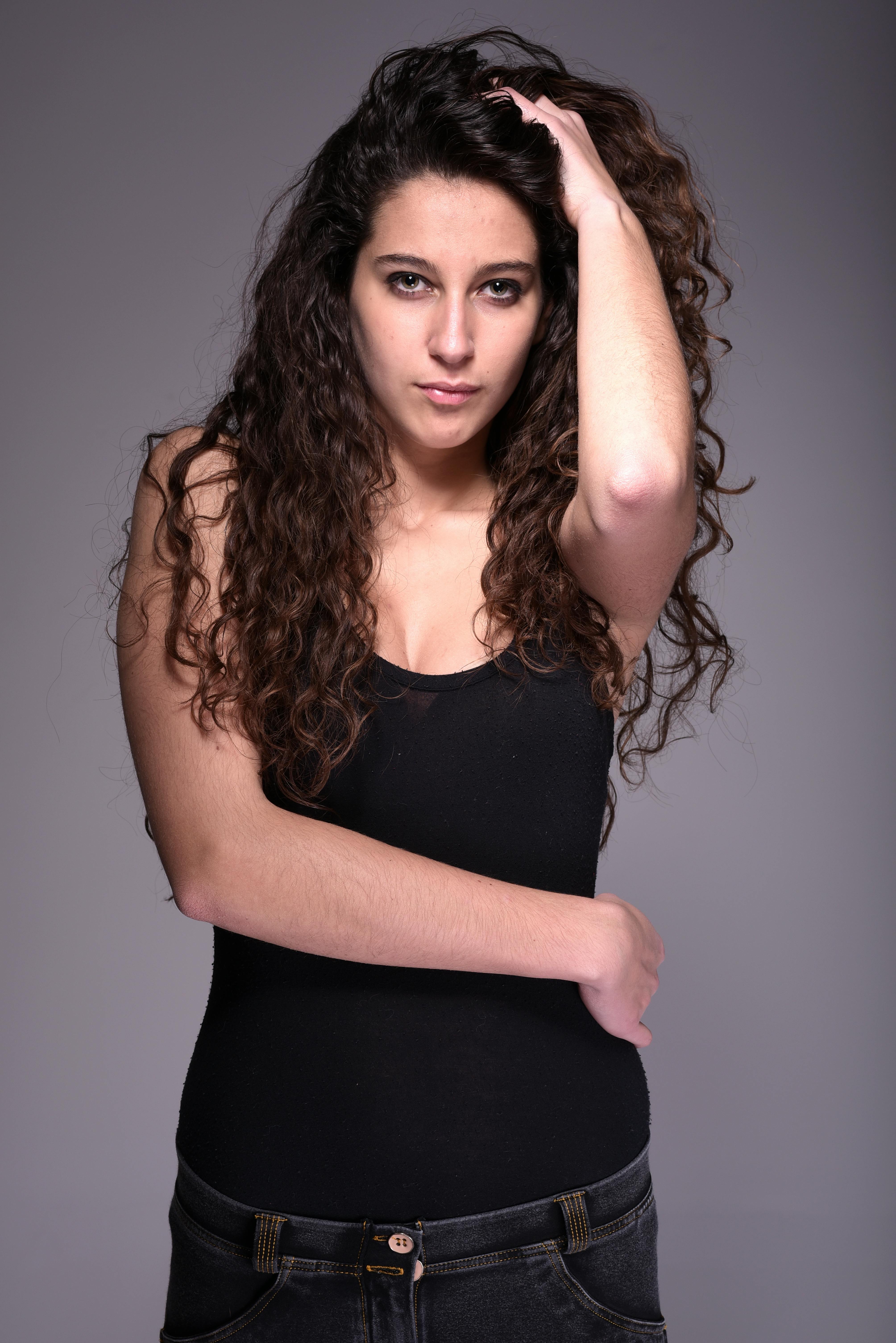 A confident young woman in a black tank top poses with curly hair, exuding assertiveness.