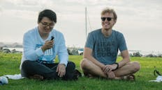 两个朋友笑容满面地坐在水边的草地上，放松身心，享受着休闲时光