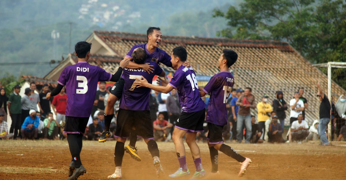 Football players celebrating a goal in a dusty outdoor match with spectators.