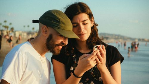 Free A couple sharing a moment on the beach during sunset, looking at a smartphone. Stock Photo