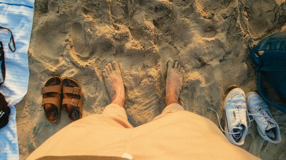 Person standing barefoot on a sandy beach with sandals and sneakers visible.
