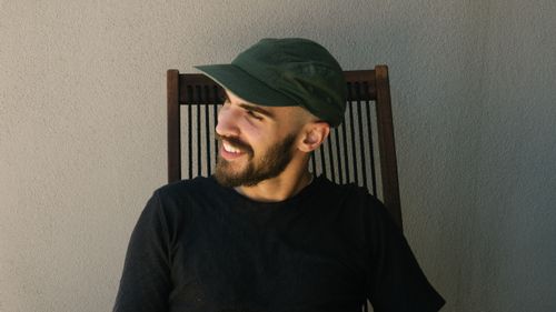 Free A relaxed man with facial hair and a cap is smiling softly while sitting outdoors, enjoying the moment. Stock Photo