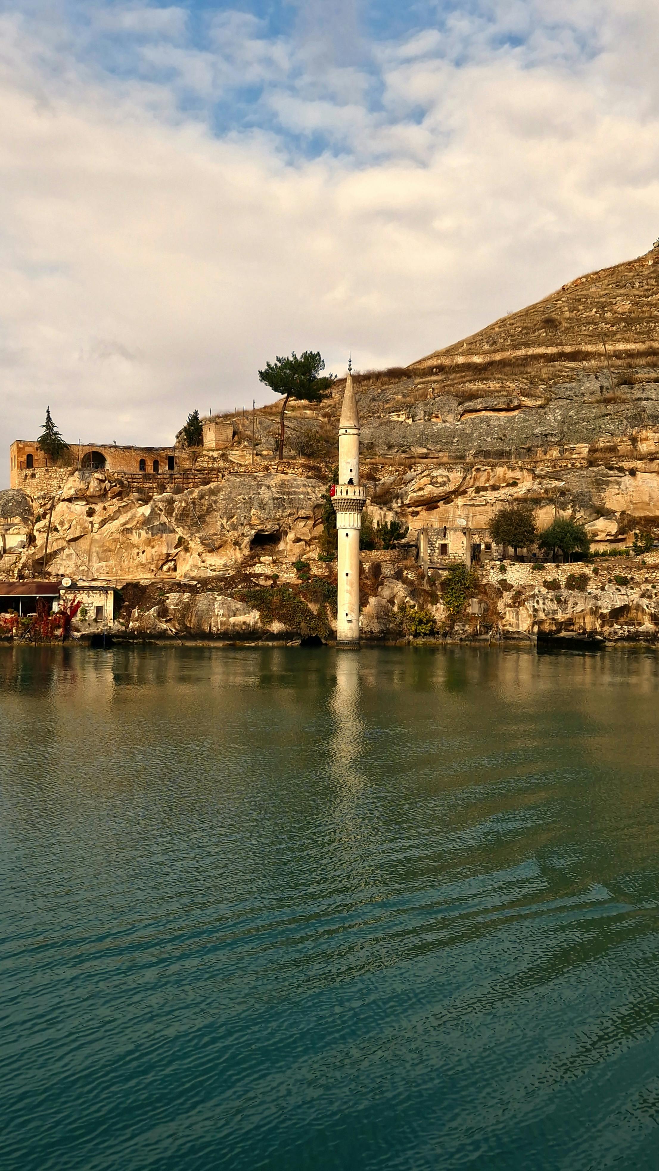 Minaret rising from waters, set against ancient rocky cliffs under a cloudy sky.