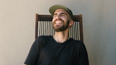 一个留着胡须、面带笑容的男人，头戴帽子，随意地坐在户外，散发着欢乐与放松的气息