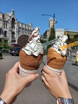 Two hands hold tasty ice cream chimney cones with toppings on a sunny city street.