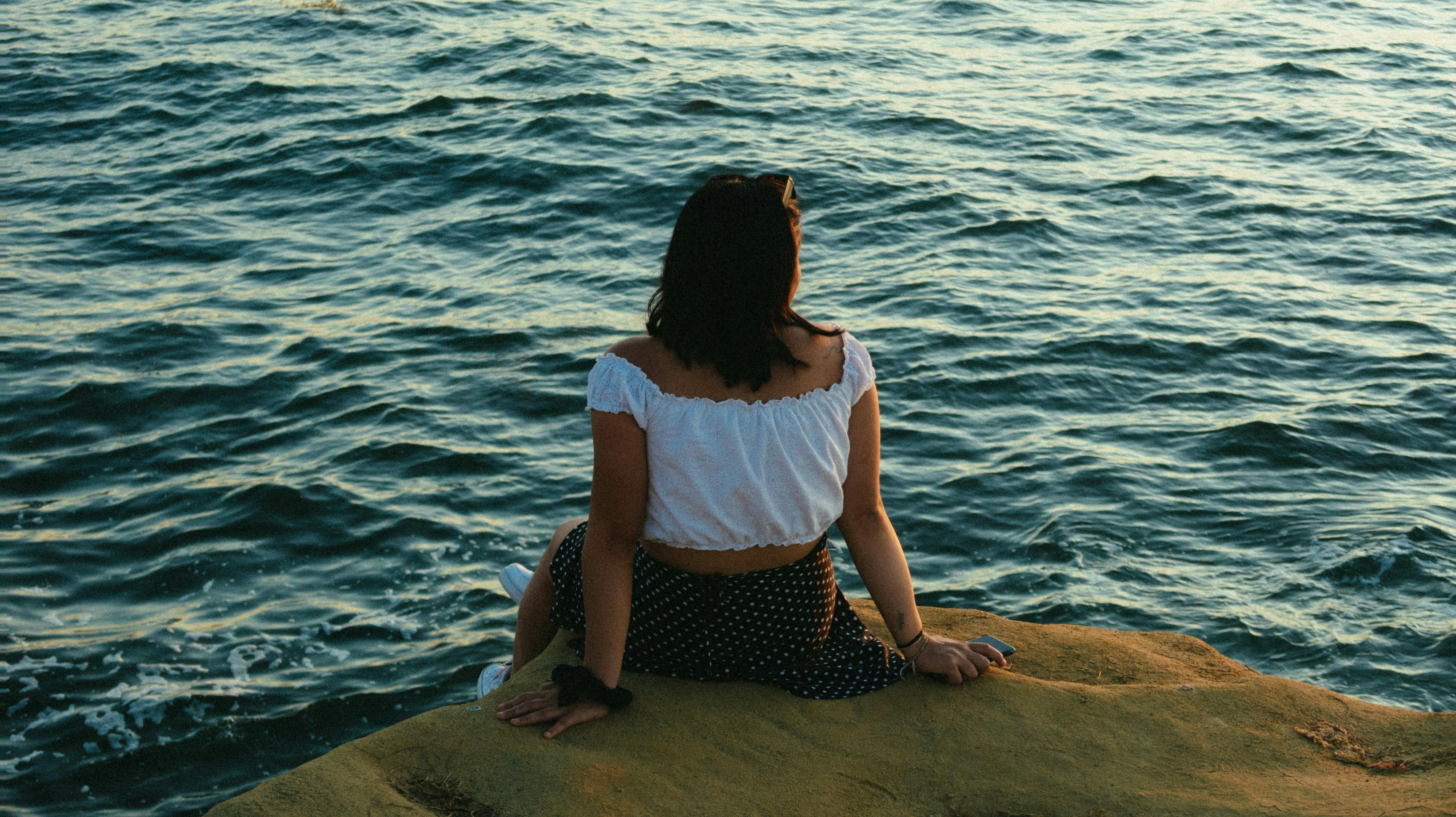 一名身着夏装的女子坐在海边的岩石上，倒映着日落