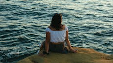 一名身着夏装的女子坐在海边的岩石上，倒映着日落