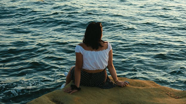 一名身着夏装的女子坐在海边的岩石上，倒映着日落