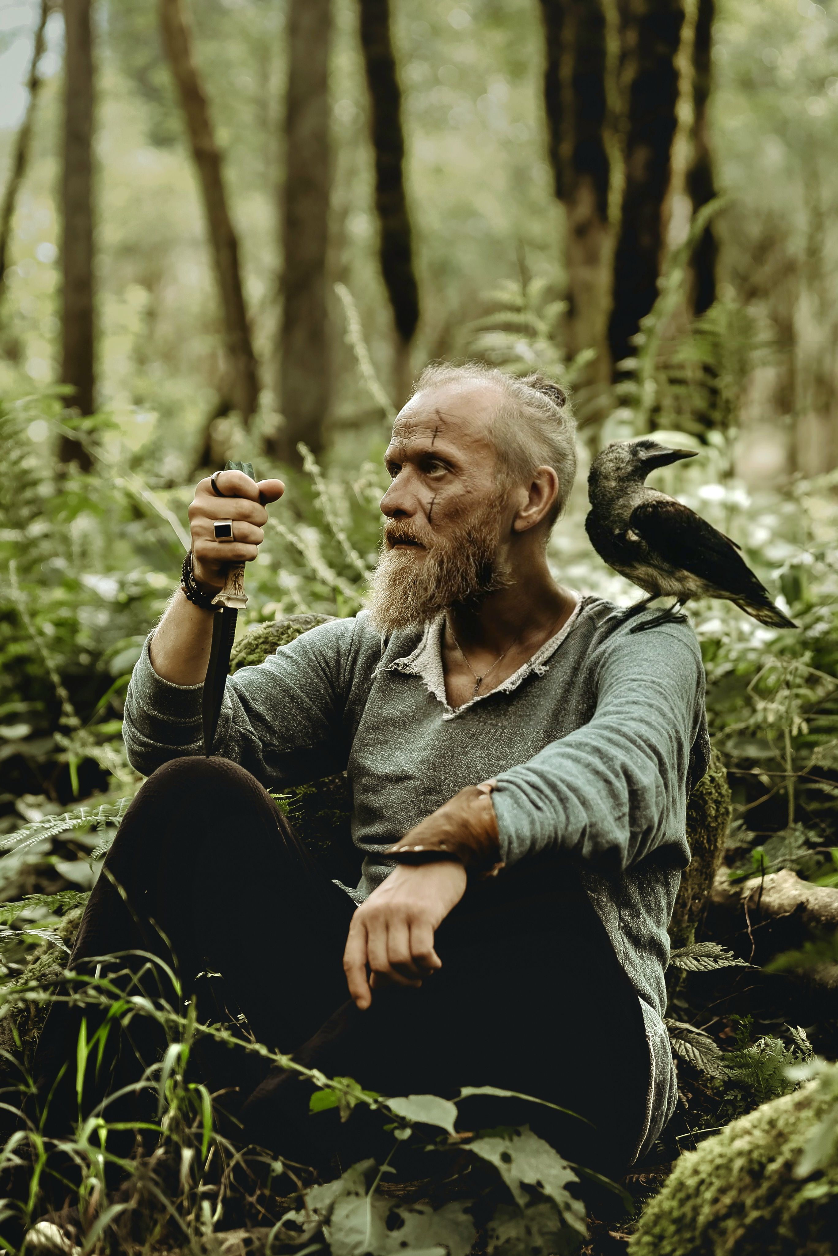 A senior man sitting in a lush forest with a crow on his shoulder, creating a mystical vibe.