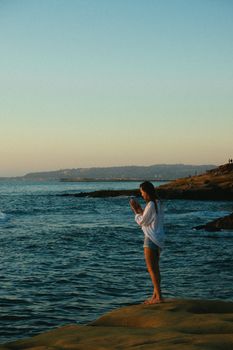 Woman stands on a rocky beach at sunset, capturing photos of the serene ocean view, enjoying leisure moments.