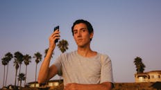 日落时分，一名年轻人在棕榈树的背景下，于户外举起了智能手机