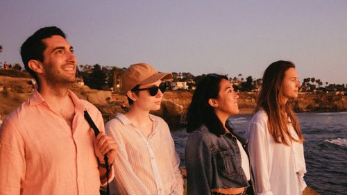 Free A group of friends smiling and enjoying a serene sunset by the sea, capturing togetherness and happiness. Stock Photo