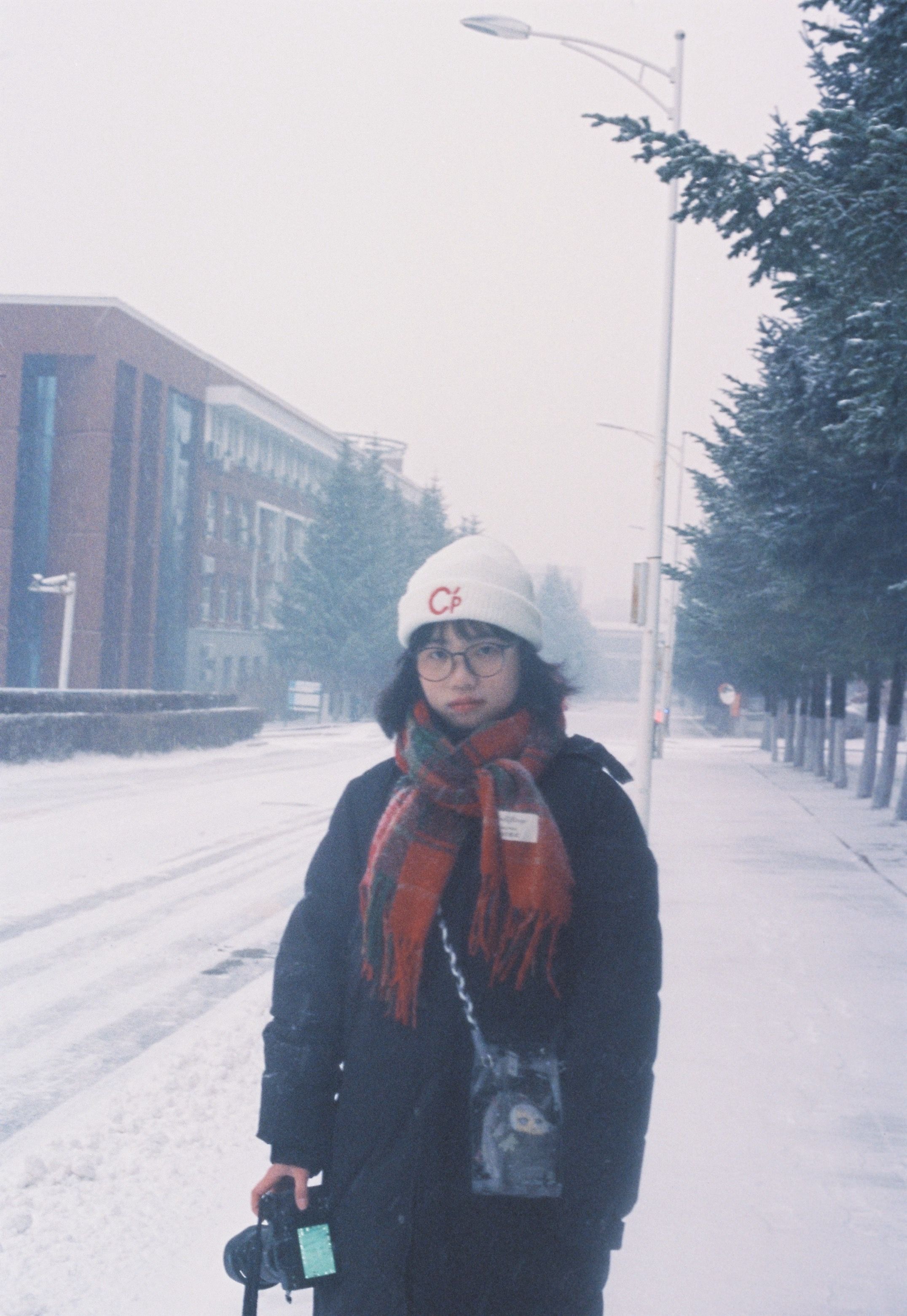 A young woman in winter attire stands on a snow-covered street, surrounded by trees and buildings.