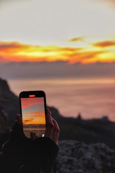 Hands holding a smartphone capturing a vivid ocean sunset, showcasing warm colors.