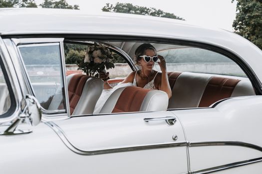 Bride sitting in a vintage car, wearing heart-shaped sunglasses and holding a bouquet. Perfect for wedding themes.