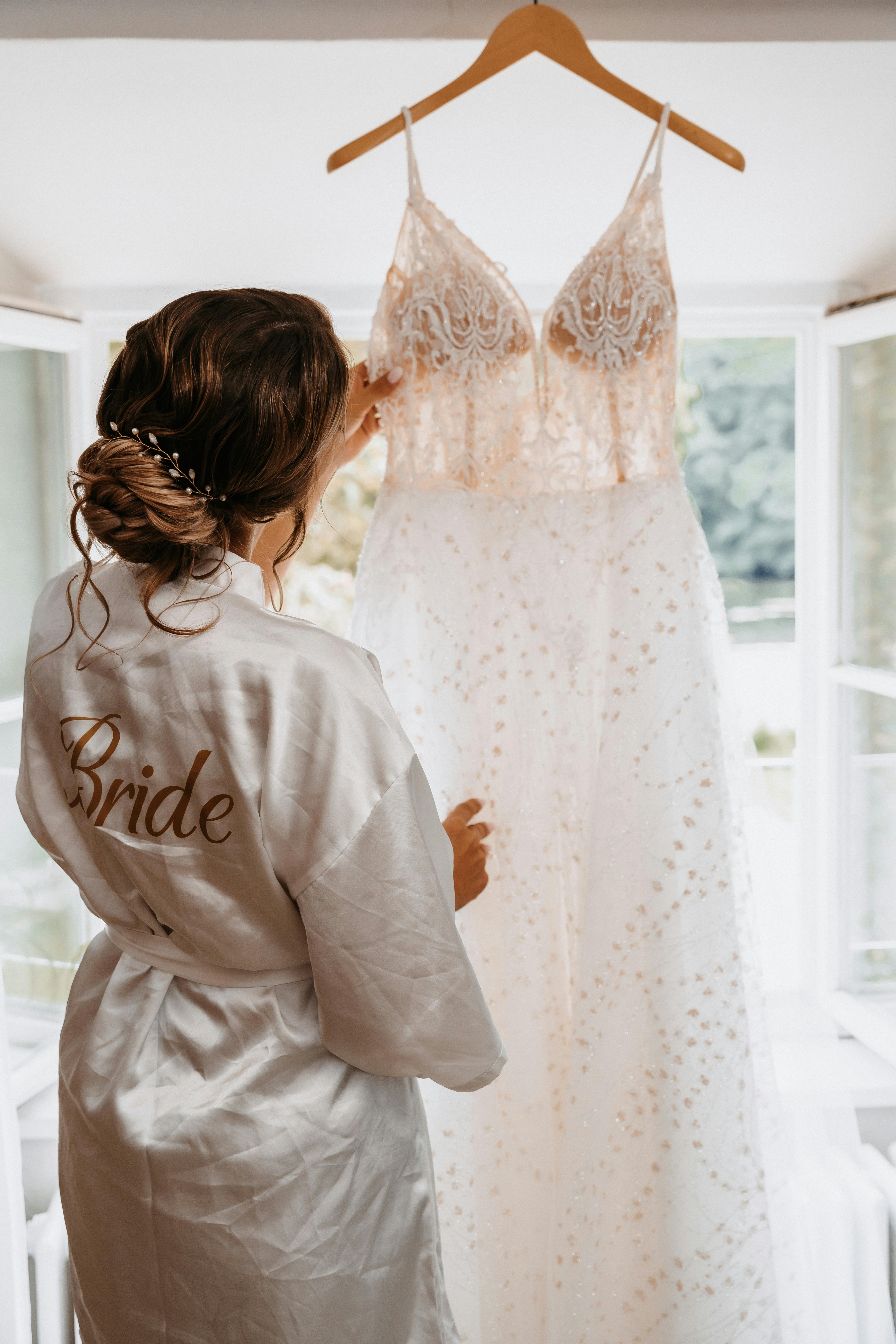 A bride in a robe admires her wedding dress hanging by a window.