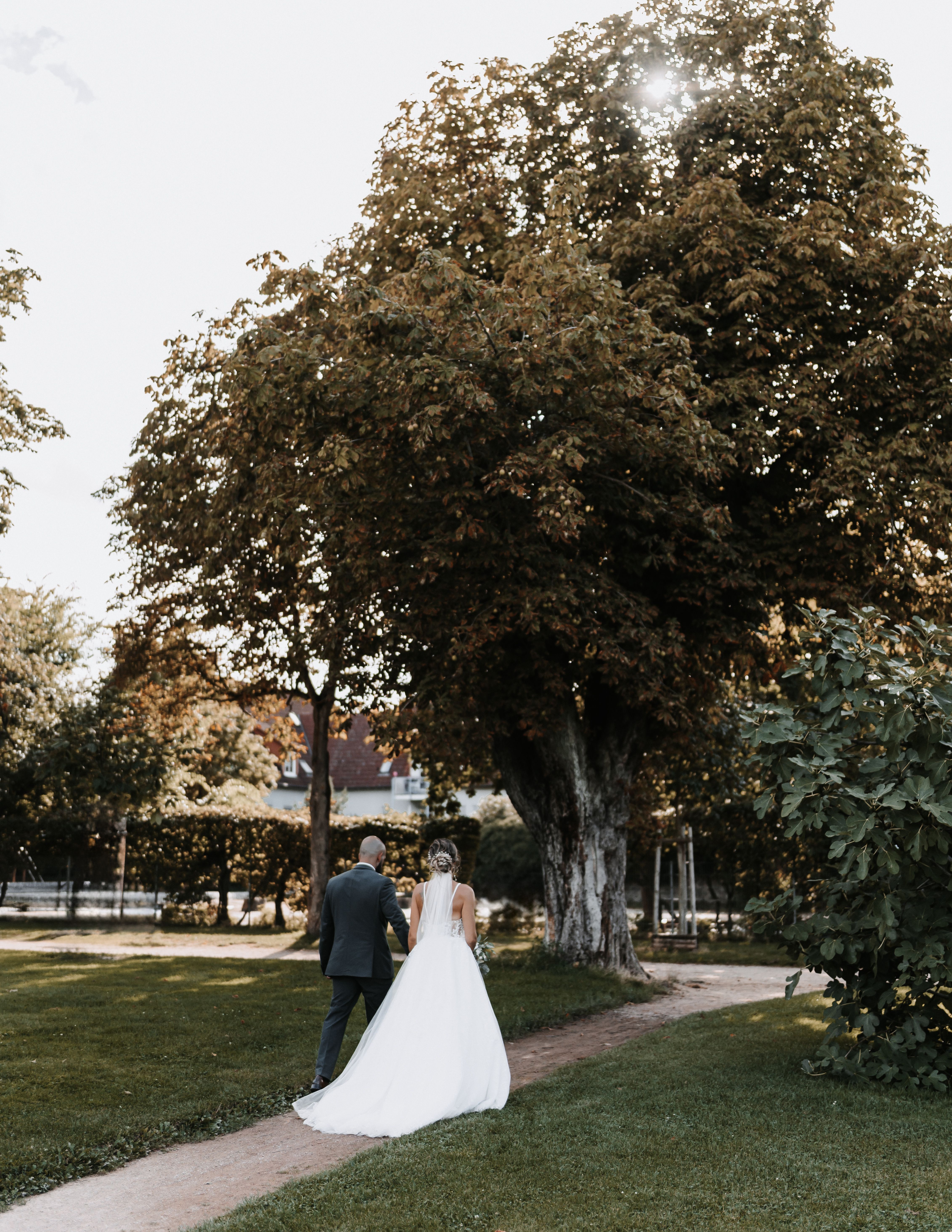 Bride and groom walking down a scenic path under a large tree in a sunny park.