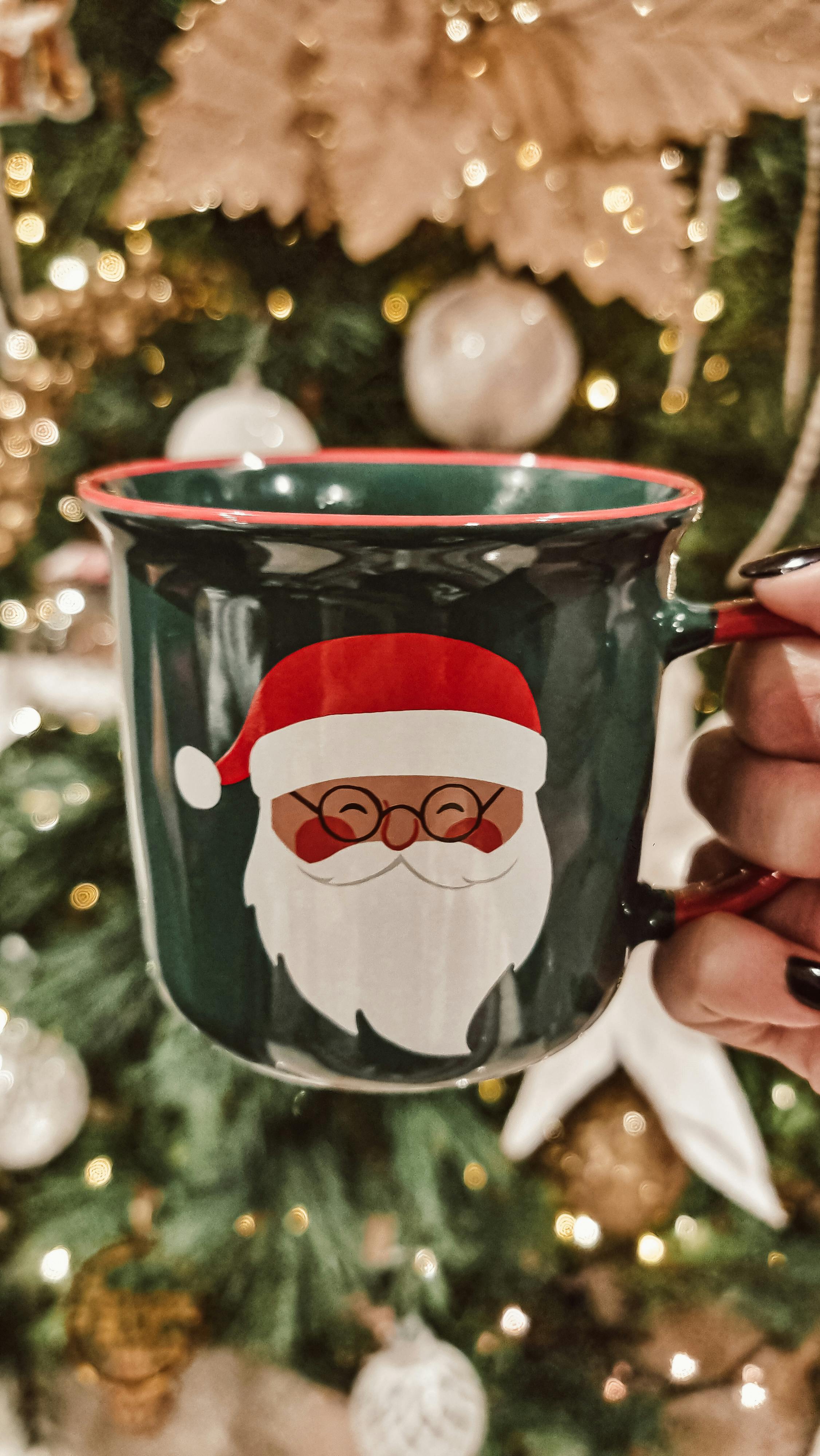 Green mug with Santa Claus design held in front of decorated Christmas tree.