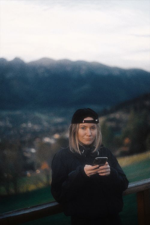 Free A young woman wearing a cap uses her phone outside with a mountain view backdrop. Stock Photo