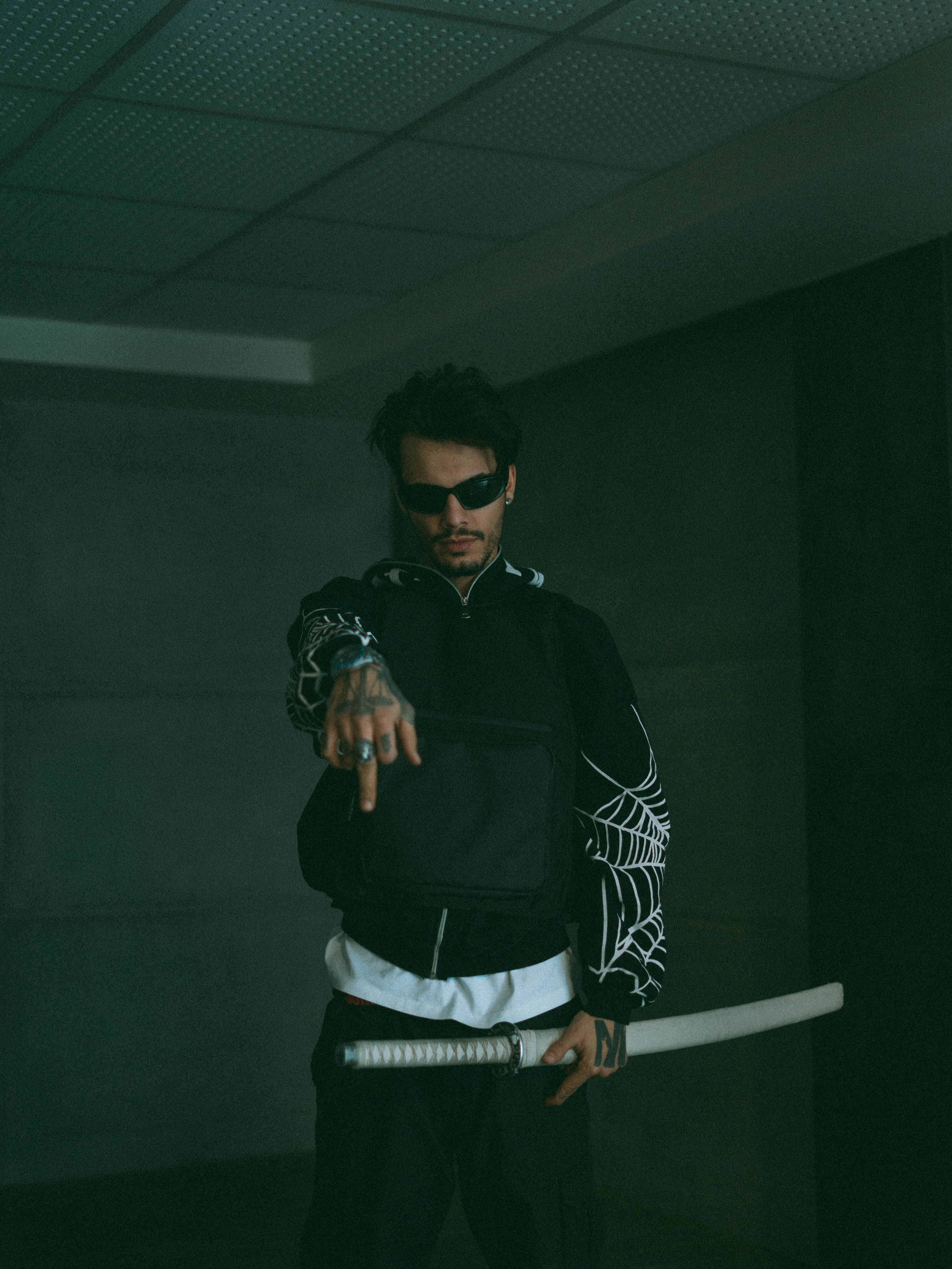 A man wearing sunglasses in a dimly lit room holding a samurai sword, featuring tattoos and casual attire.