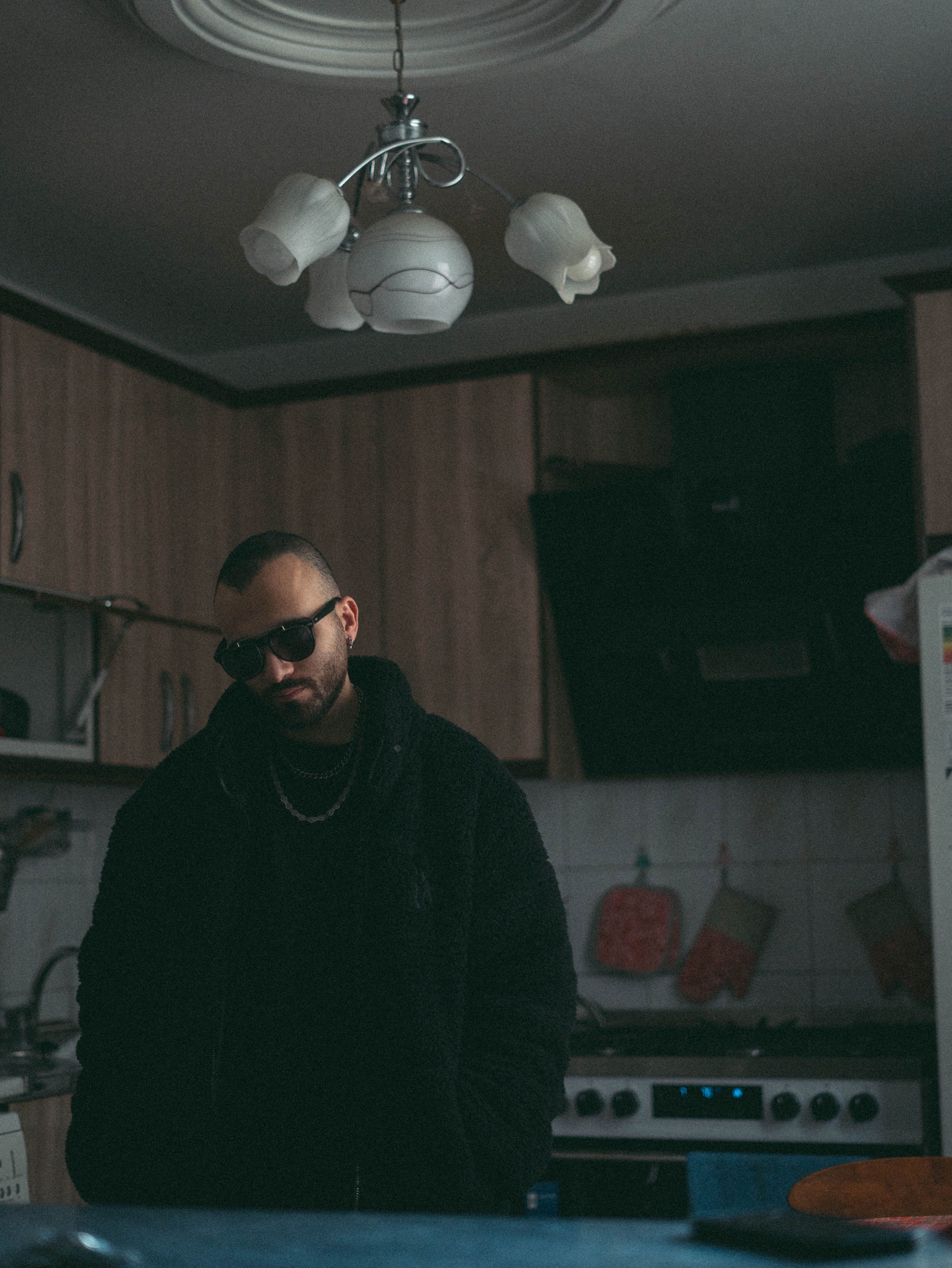 A man in sunglasses stands in a dimly lit kitchen, exuding a mysterious vibe.