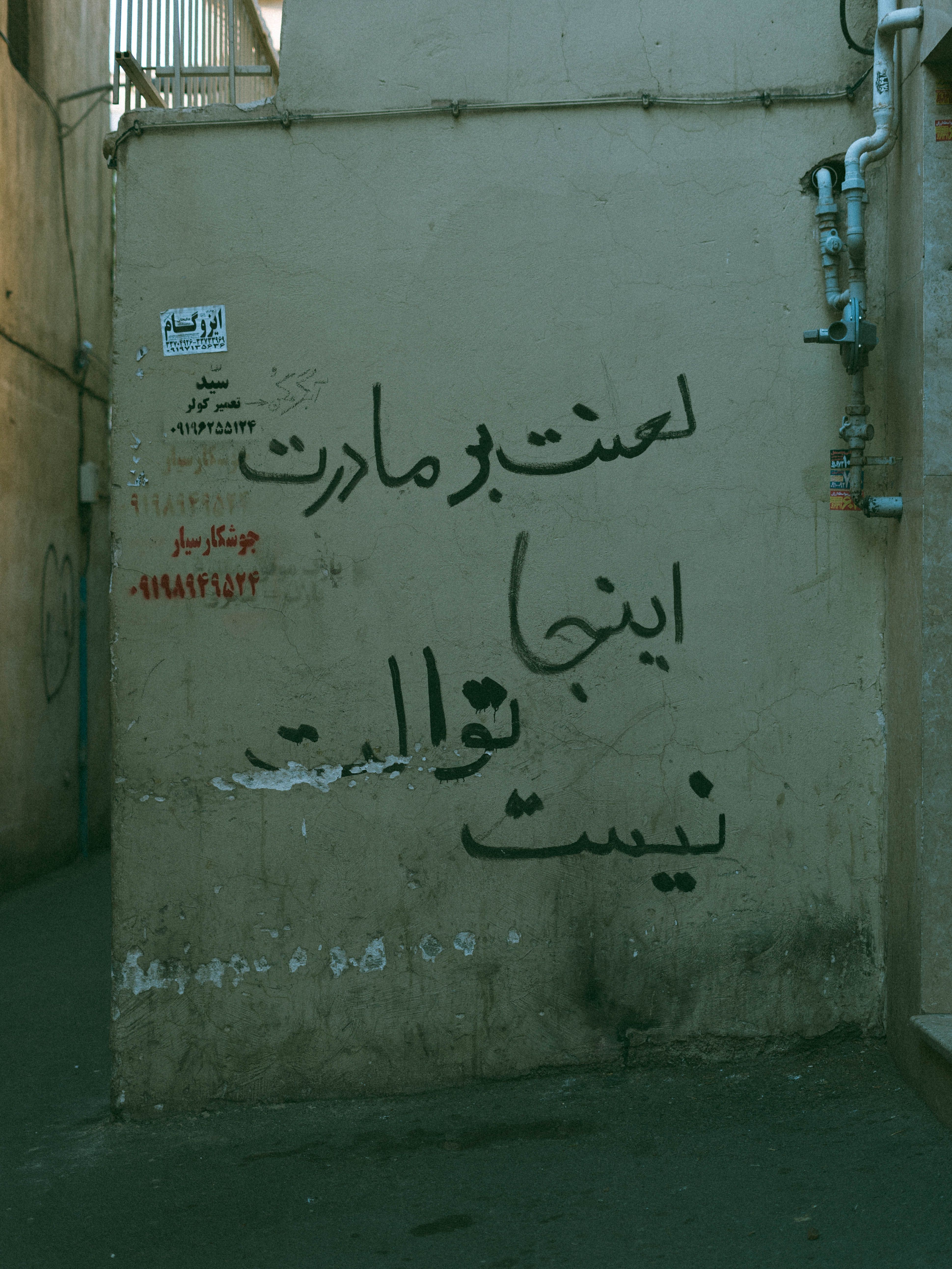 Graffiti text on a concrete wall in an urban alley, featuring pipes and electrical boxes.
