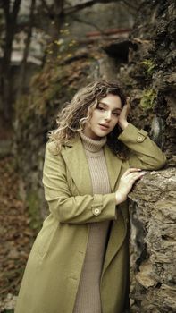 Young woman in a green coat poses thoughtfully against a stone wall in a serene outdoor setting.