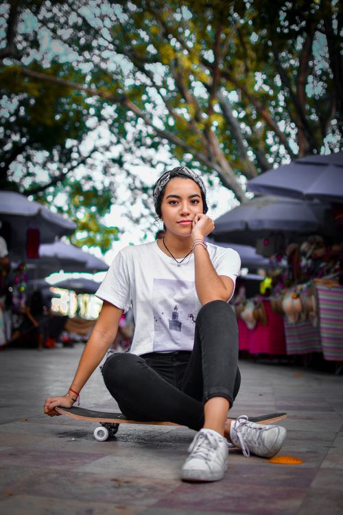 Free Casual young adult woman sitting with a skateboard in a vibrant outdoor market, expressing relaxed urban style. Stock Photo