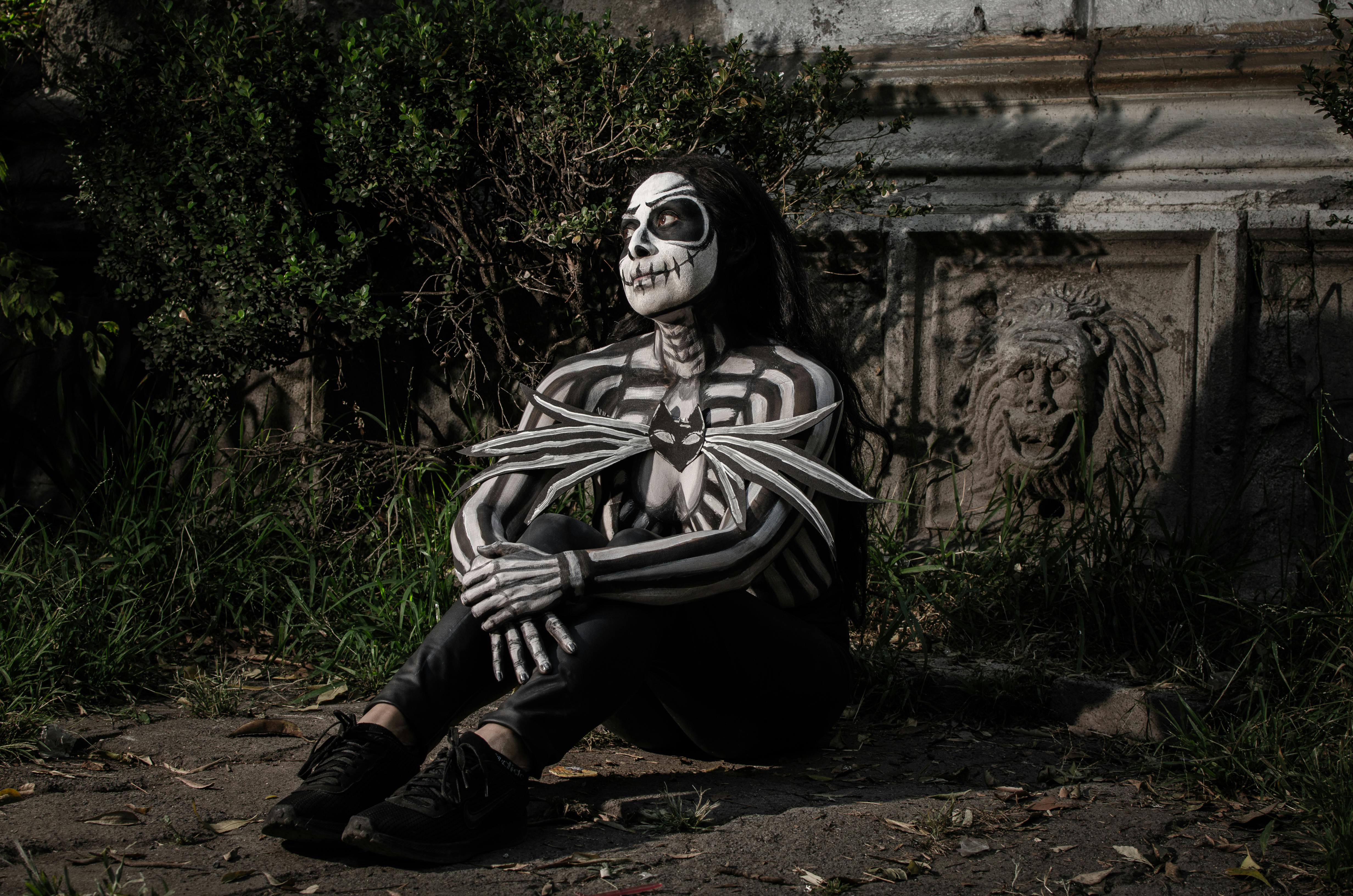 Person in skeletal body paint sits in a gothic outdoor setting, evoking Halloween vibes.