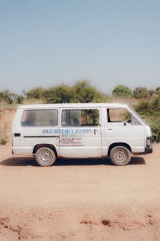 White minibus parked on a dusty road in Katsina, Nigeria, under a clear sky.