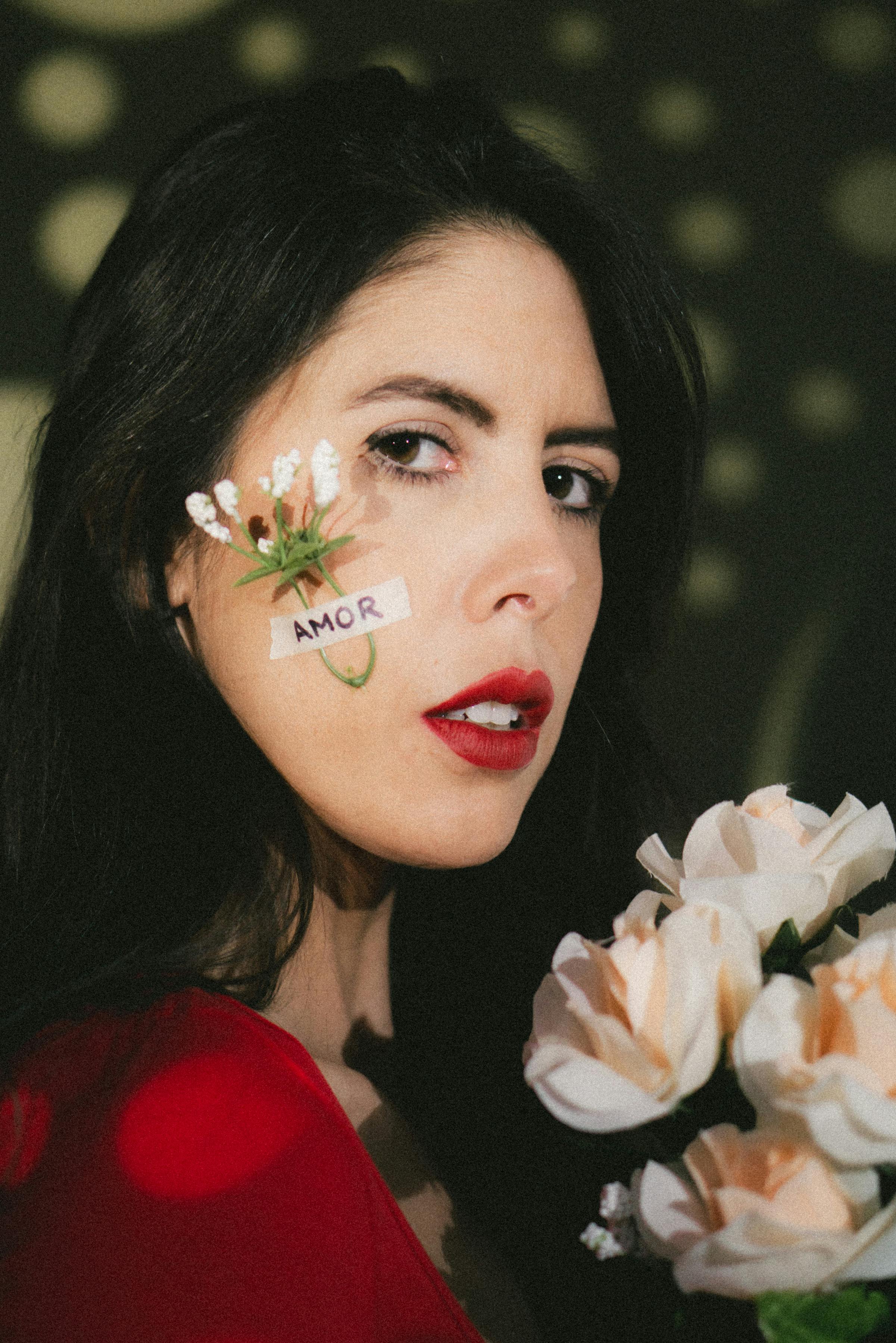 A portrait of a woman with small flowers and 'amor' sticker on her cheek, holding roses.