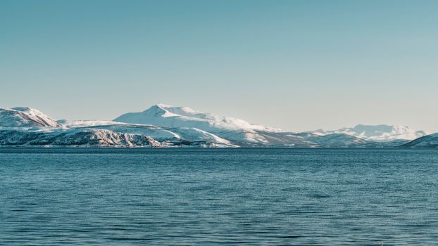 Scenic view of snow-covered mountains by a peaceful ocean under blue skies in Northern Norway.