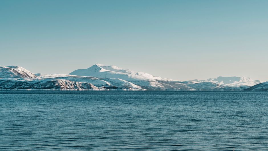 Scenic view of snow-covered mountains by a peaceful ocean under blue skies in Northern Norway.