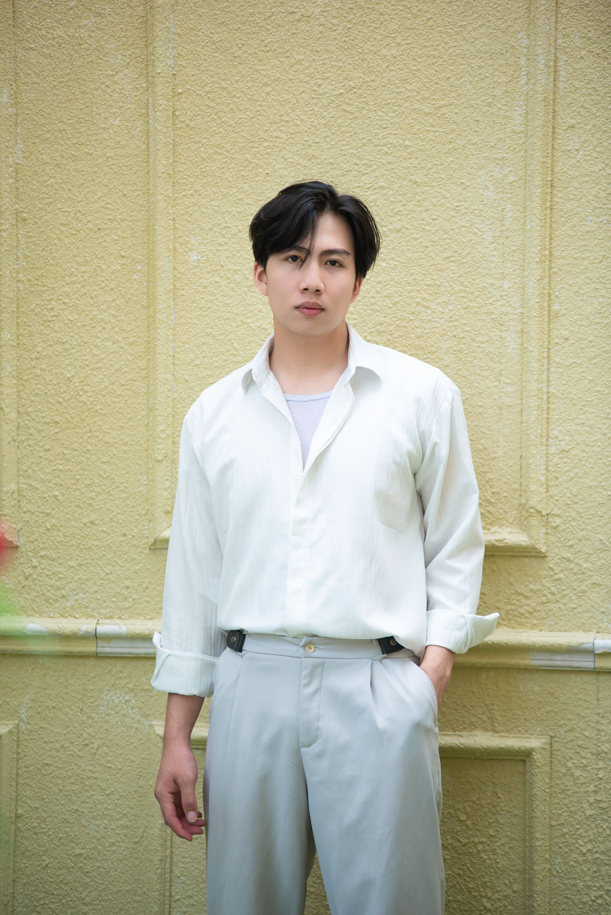 Stylish young man in white shirt posing against a textured wall outdoors.