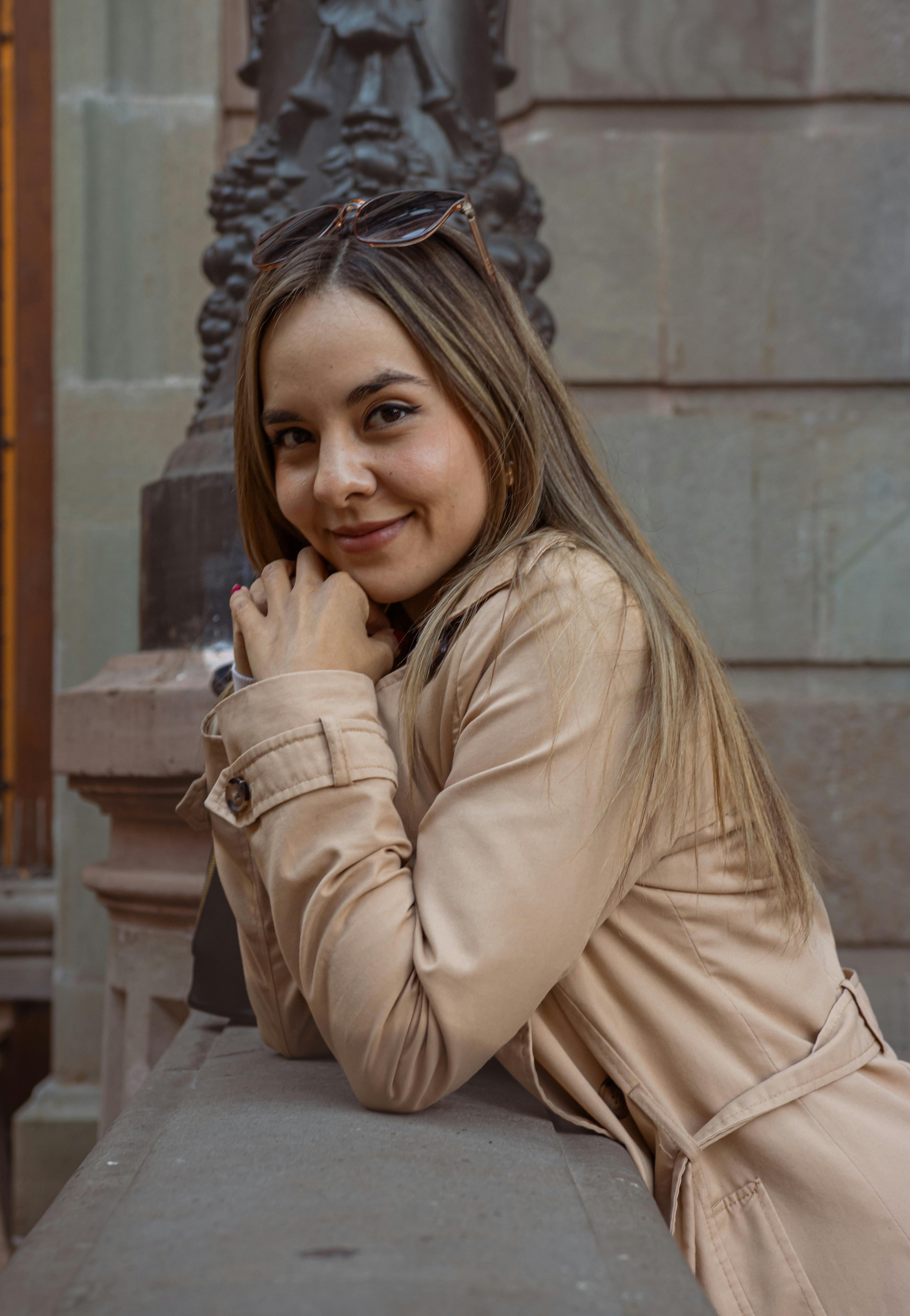 Ücretsiz Genç bir kadın dışarıda bir korkuluğa yaslanmış, şık bej bir trençkot giymiş ve sıcak bir şekilde gülümsüyor. Stok Fotoğraflar