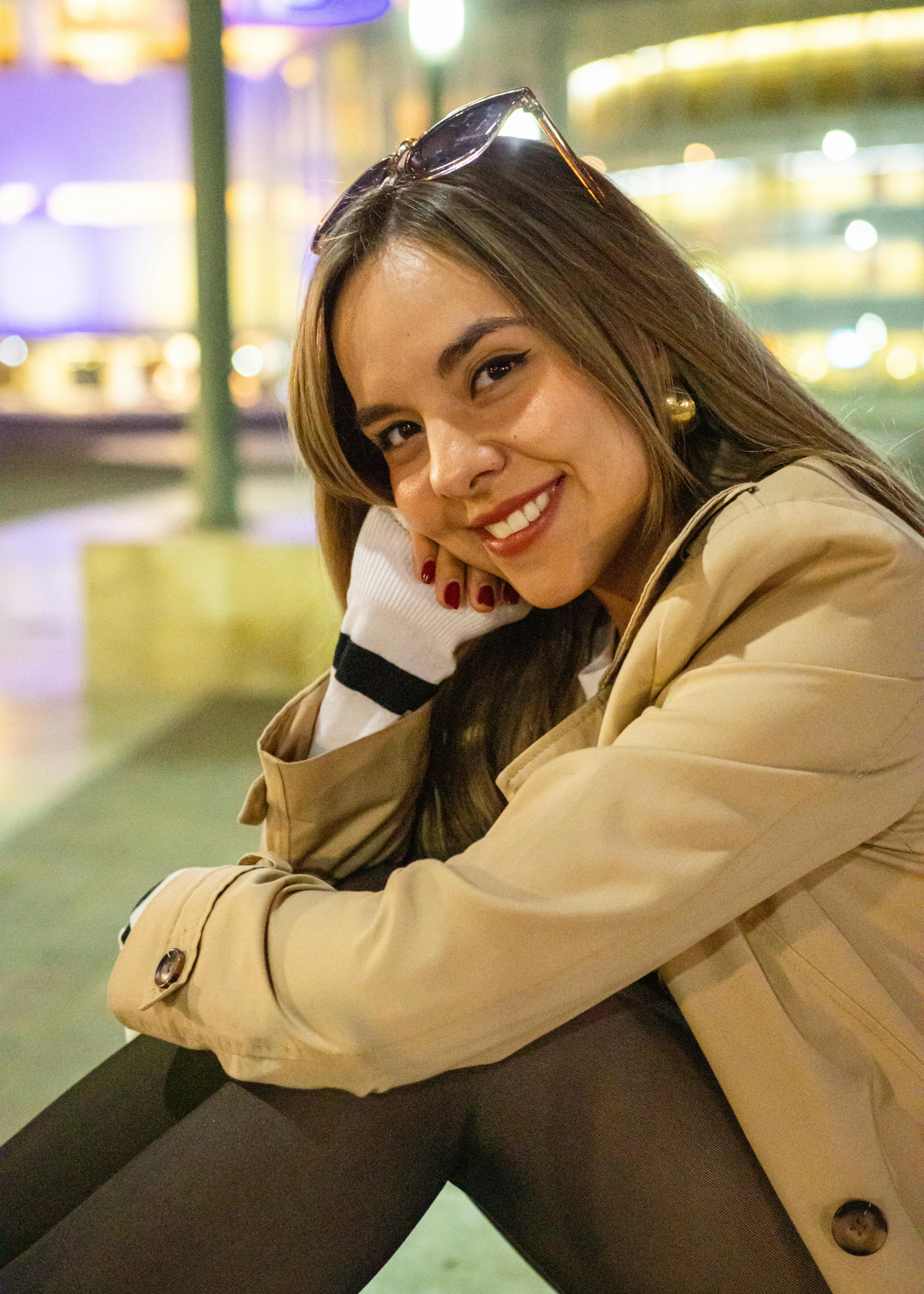 Free Portrait of a young smiling woman in a coat with urban lights in the background. Stock Photo