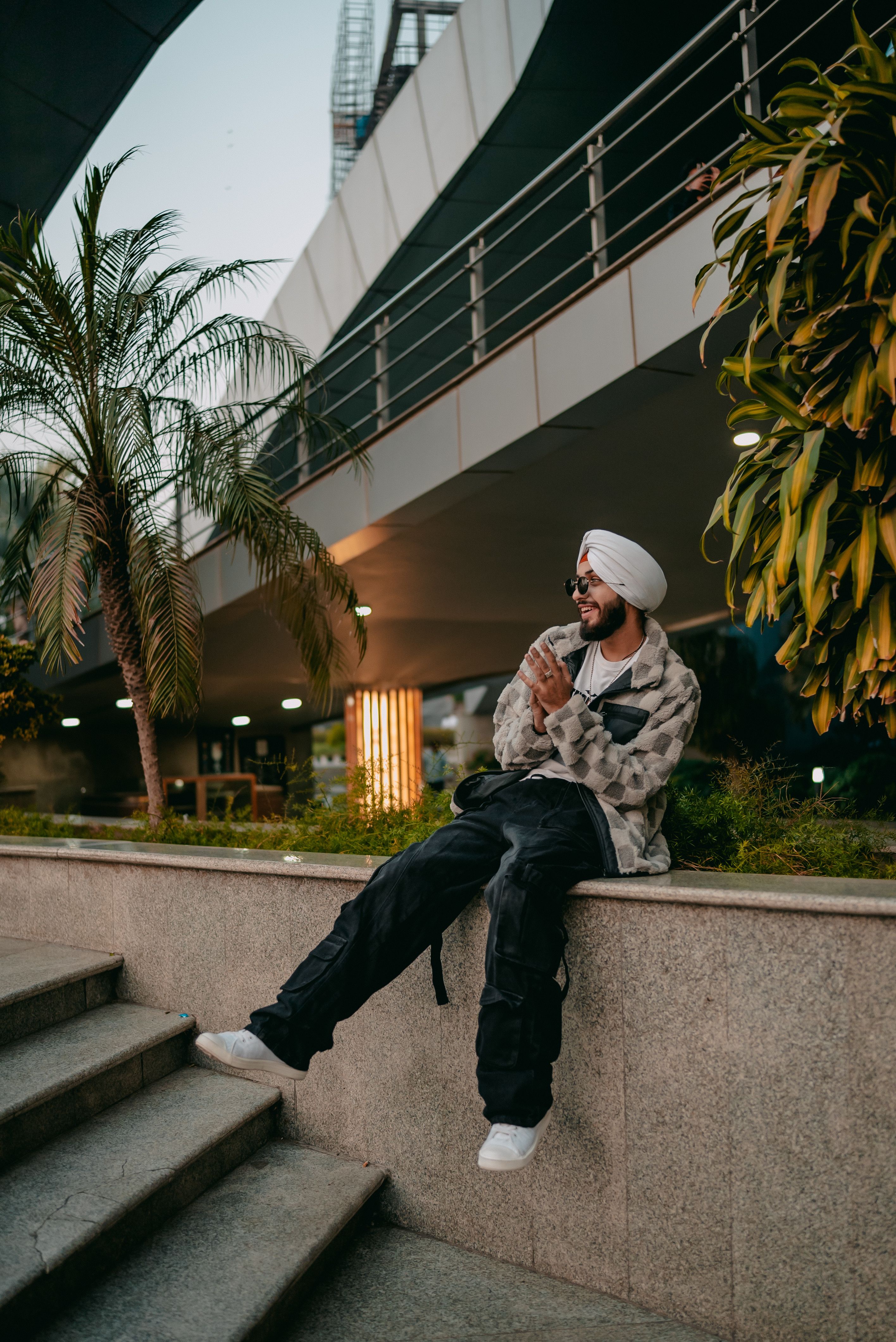 A man wearing a turban sits casually outside a modern architecture building, enjoying the moment.