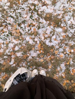 Feet in white sneakers on ground with autumn leaves and patchy snow.