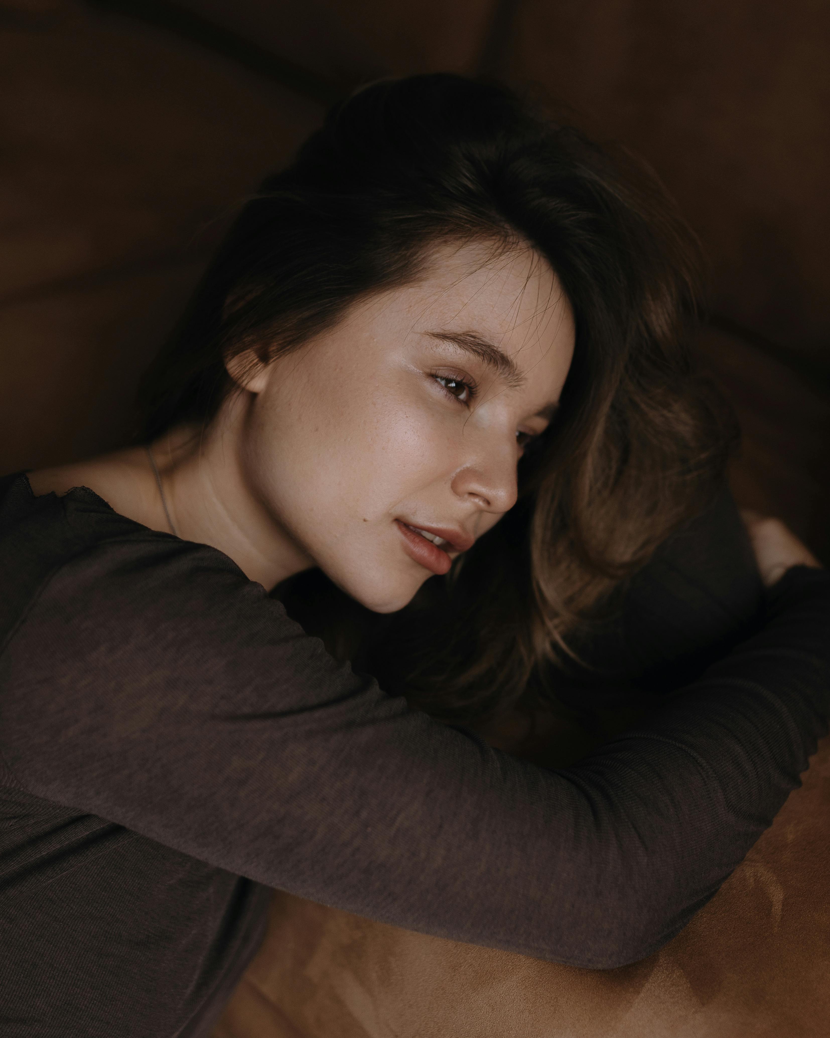 Serene woman resting in soft bedroom lighting, eyes closed in deep relaxation, face peaceful and dreamy amid gentle shadows
