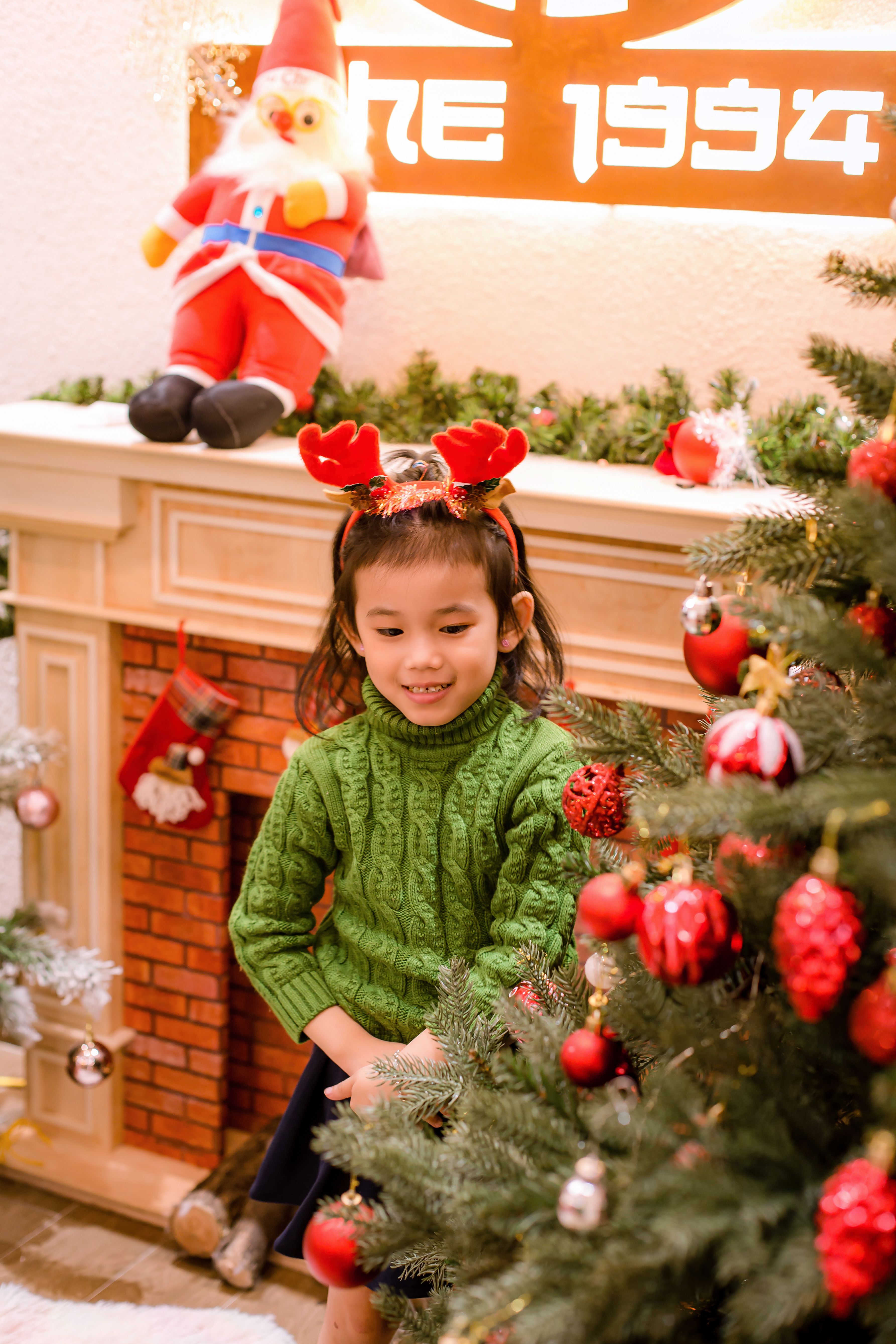 A joyful child with reindeer antlers hugging a Christmas tree indoors, Hội An.