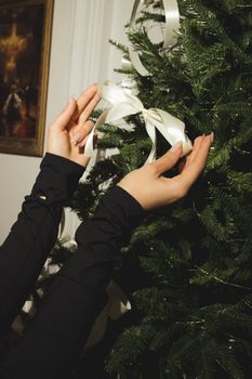 Hands decorating a Christmas tree with satin white ribbon indoors.