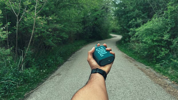 Man holding JBL speaker on a scenic forest path, perfect for outdoor music adventures.